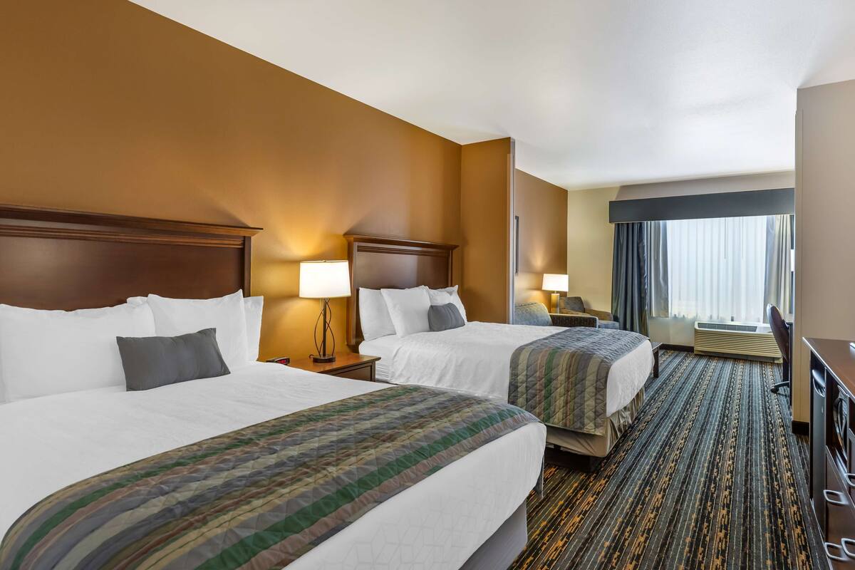 A hotel room with two beds, a large window with curtains, a desk, and a carpeted floor.