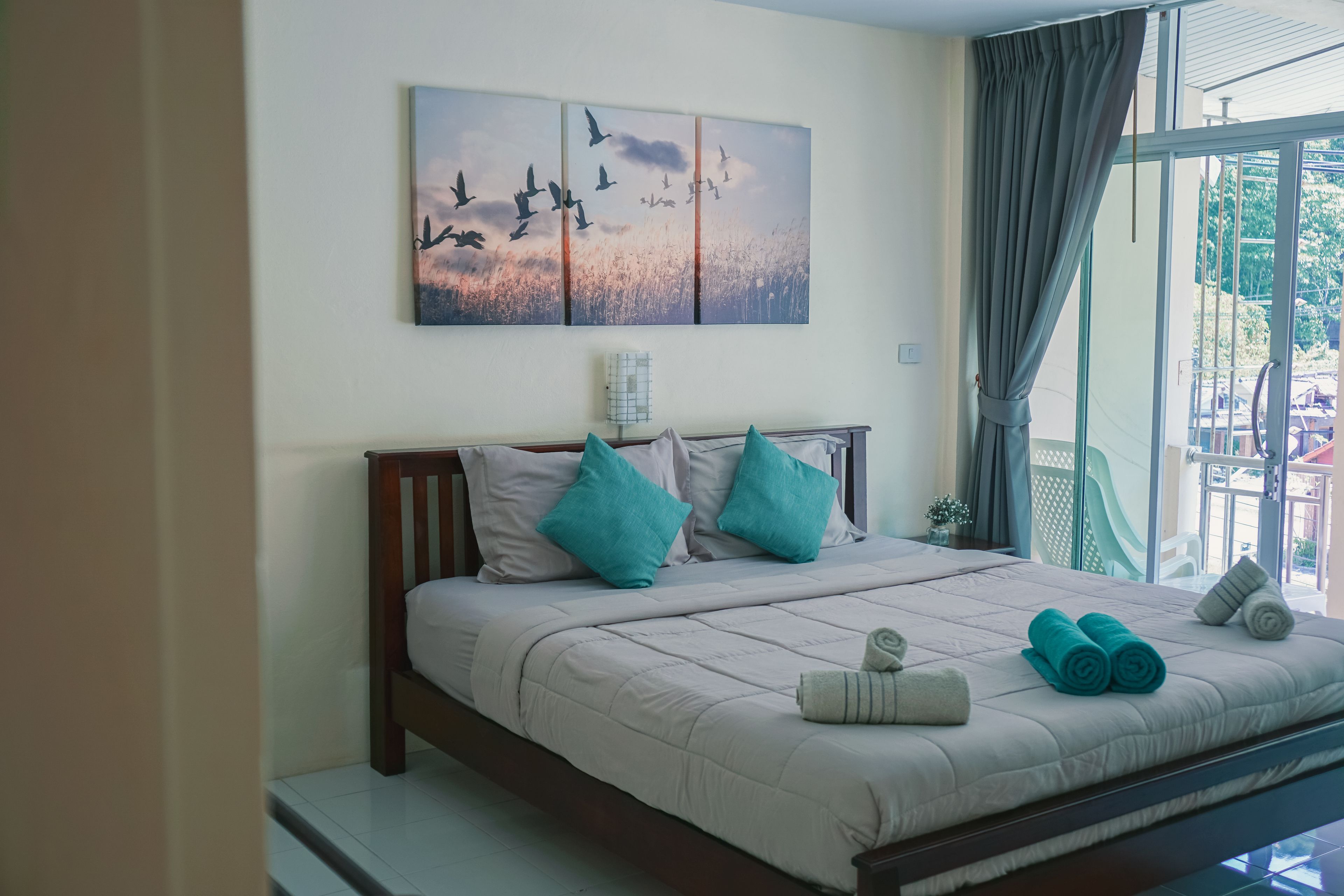A neatly made bed with a beige comforter, two teal pillows, and rolled towels. A large window with curtains is visible, and a painting of birds in flight hangs on the wall.