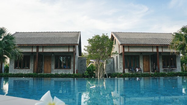 Vista de la piscina en un resort, con dos edificios, una pileta transparente y una flor blanca al frente.