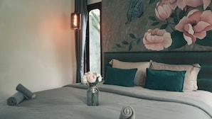 Una cama bien hecha con un florero, una toalla enrollada y vistas a un cuarto con un mural floral y una ventana.