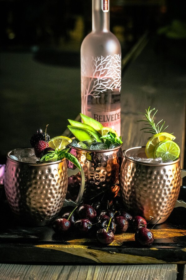Two copper mugs with ice and garnishes, a bottle of Belvedere, and cherries on a wooden surface. - Hotel Royal Palace (Hermosillo)