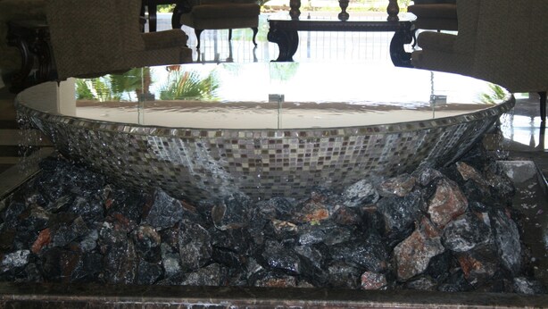 Un elemento acuático grande y circular con mosaicos, base de piedra, zonas de estar alrededor y vistas a una piscina y vegetación.