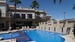 Una piscina con pared de piedra y palmeras, rodeada por un edificio con balcones y mobiliario exterior.