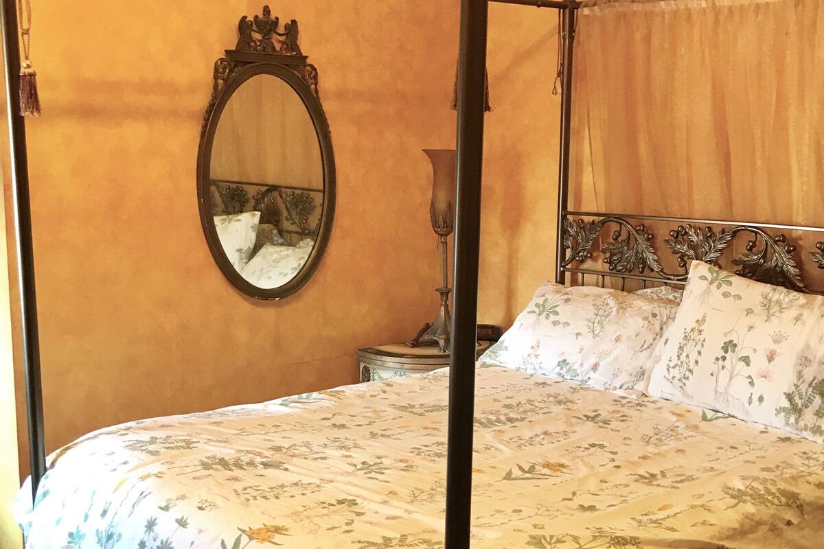 A four-poster bed with floral bedding, a round mirror with an ornate frame, and a bedside table with a lamp.