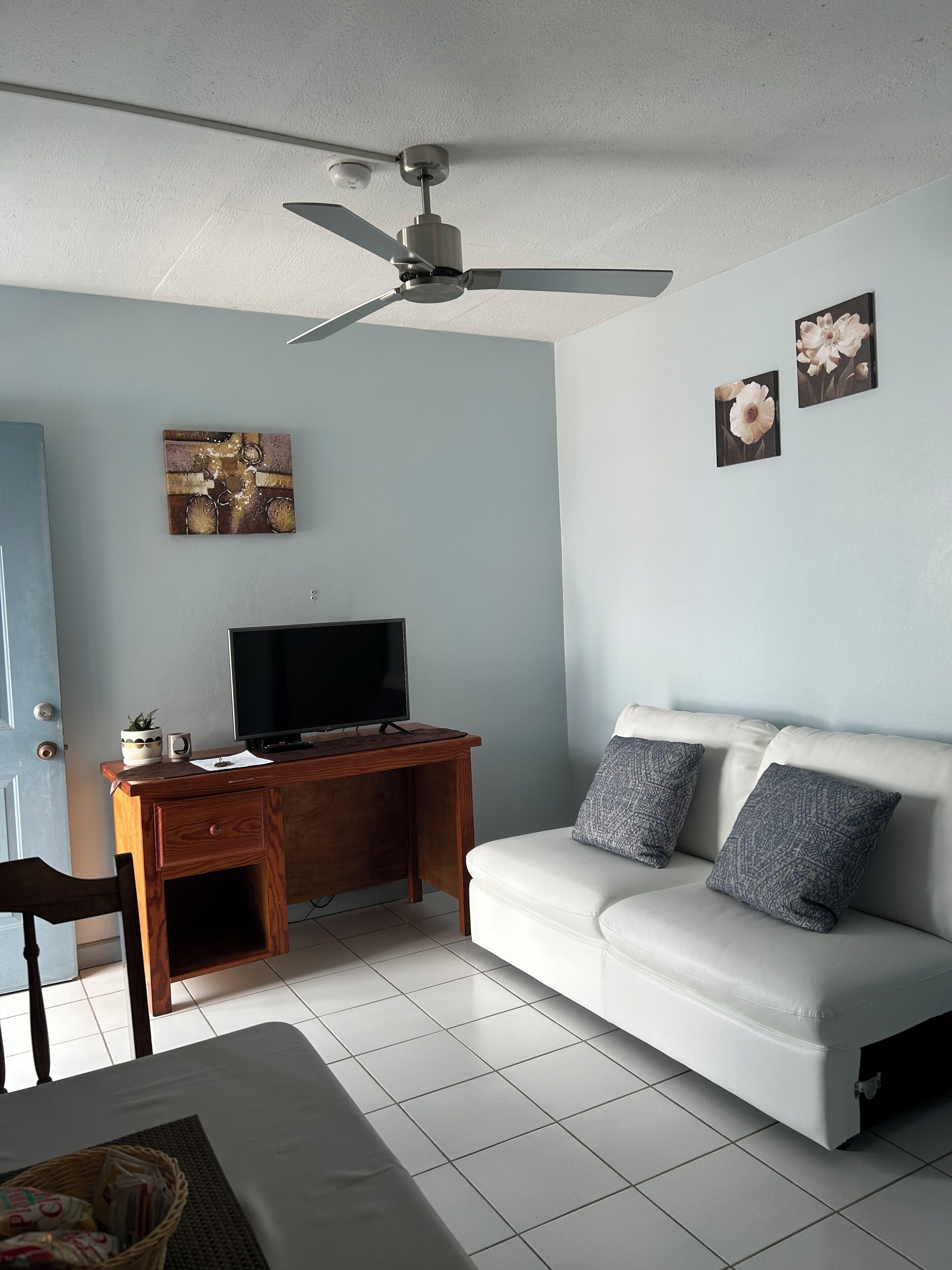 A room with a ceiling fan, a wooden desk with a TV, a white sofa with patterned cushions, and artwork on the wall.