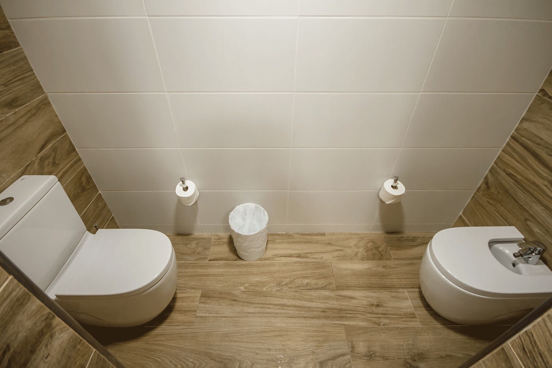 A bathroom with two toilets, a trash bin, and a toilet paper holder on the wall.