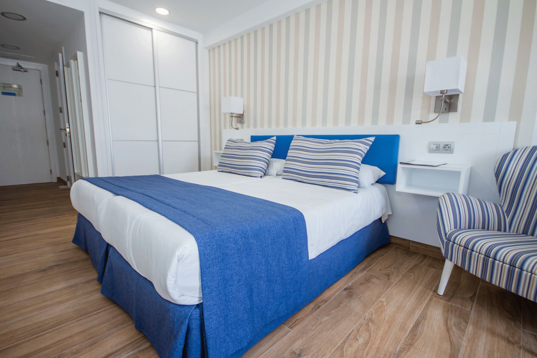Ein ordentlich eingerichtetes Hotelzimmer mit einem großen Bett, einem Nachttisch, einem Sessel und einer gestreiften Wand.