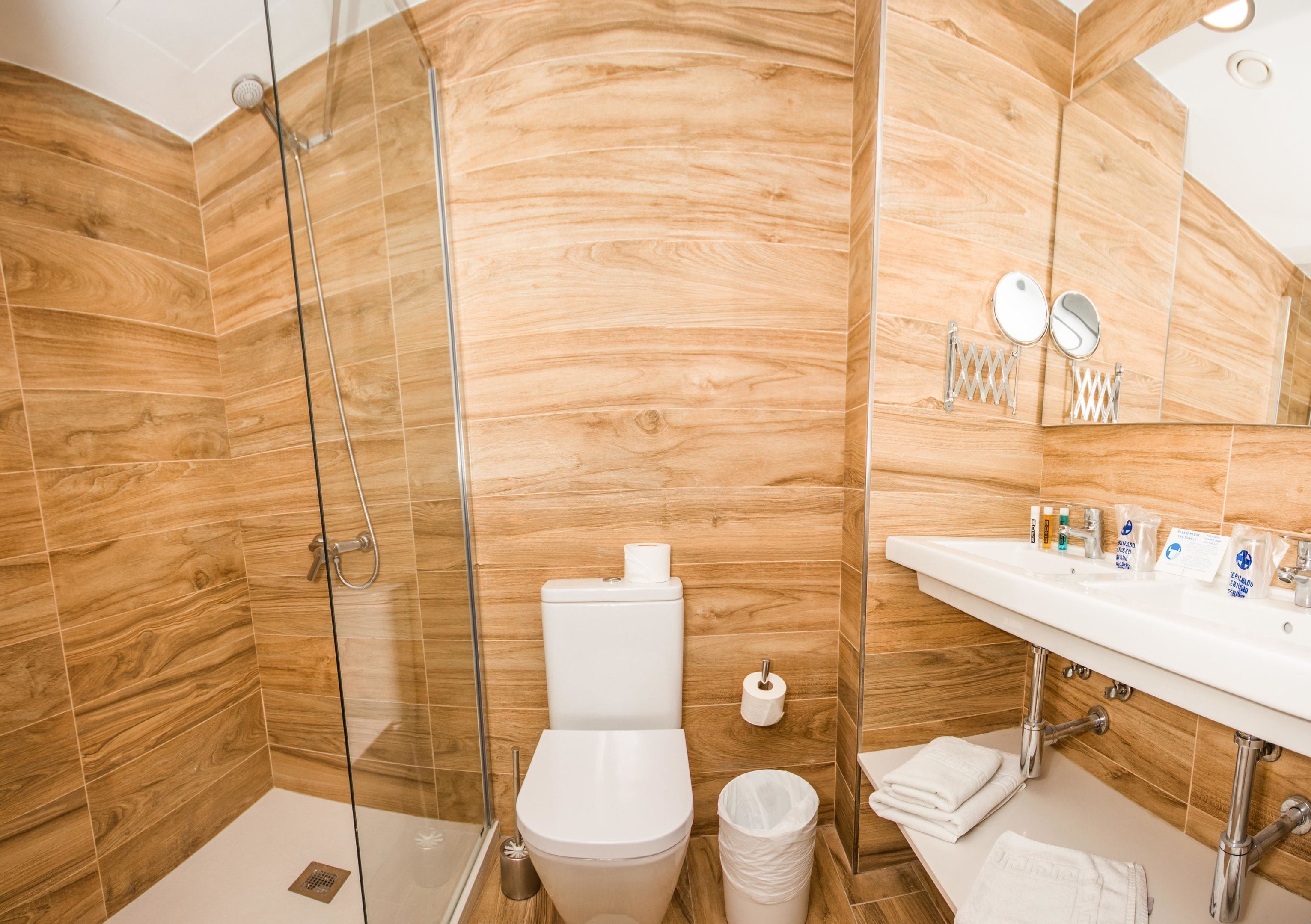 A bathroom with a wooden wall, a glass shower enclosure, a white toilet, and a sink with toiletries.
