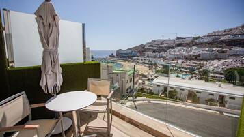 Uma varanda com mesa e cadeiras, com vista para um horizonte costeiro com edifícios e uma praia.