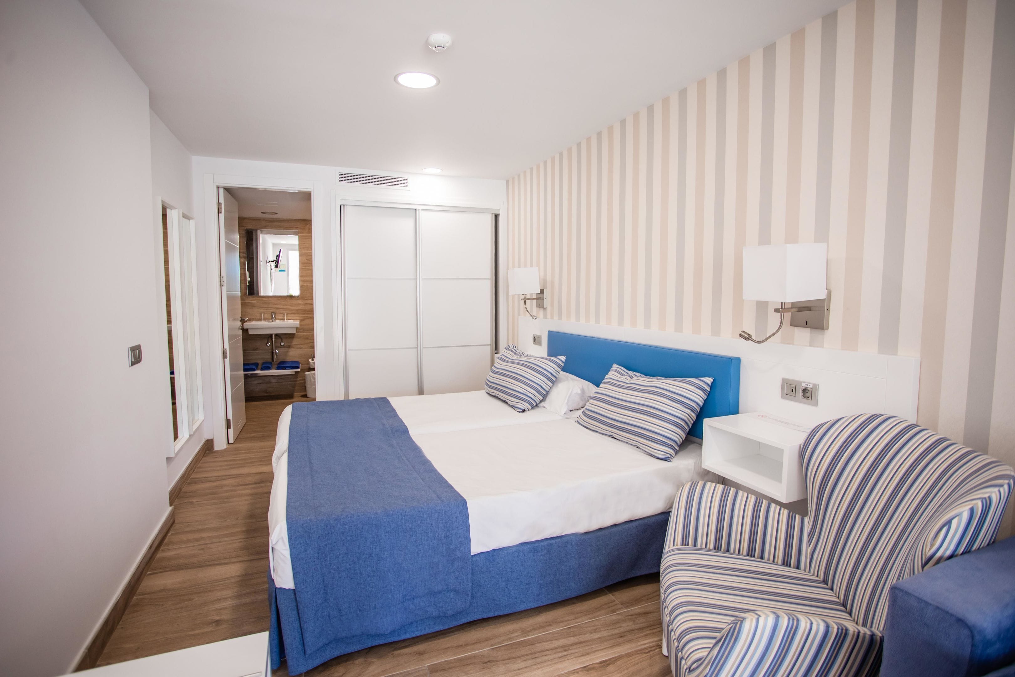 Una habitación de hotel moderna con una cama grande, una mesita de noche, una silla, un espejo y un baño visible a través de una puerta abierta.