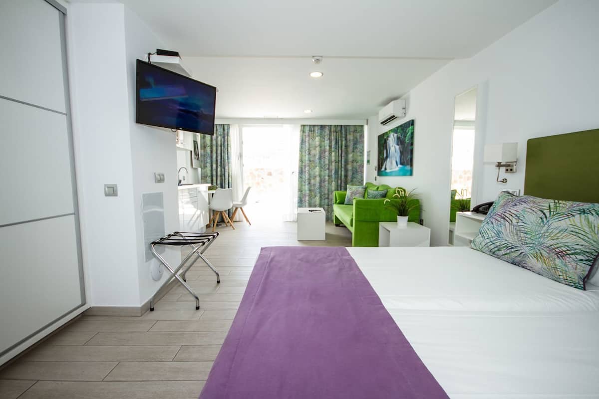 A hotel room with a bed, a desk, a green sofa, a TV, and a window with curtains.
