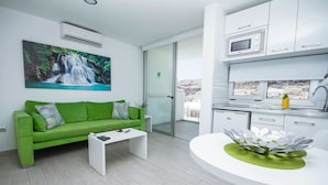 Ein modernes Wohnzimmer mit einem grünen Sofa, einem Couchtisch und einem Esstisch mit dekorativer Dekoration. Es gibt eine Küchenzeile mit weißen Schränken und einer Mikrowelle. Ein großes Fenster bietet Blick auf die Stadt.