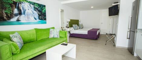 A modern hotel room with a green sofa, a bed with a purple bedspread, a white coffee table, and a painting of a waterfall on the wall.