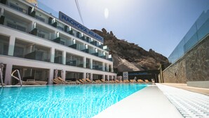 Ein Hotelpool mit klarem, blauem Wasser, Liegestühlen und einer felsigen Klippenlandschaft im Hintergrund.