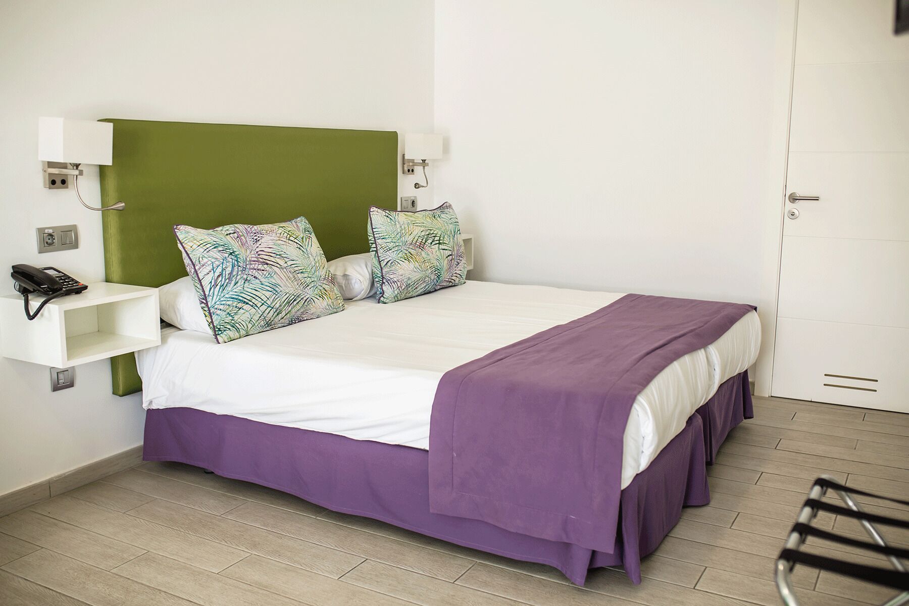 A hotel room with a green headboard, a white bedside table with a phone, two patterned pillows, a purple bedspread, and a wooden floor.