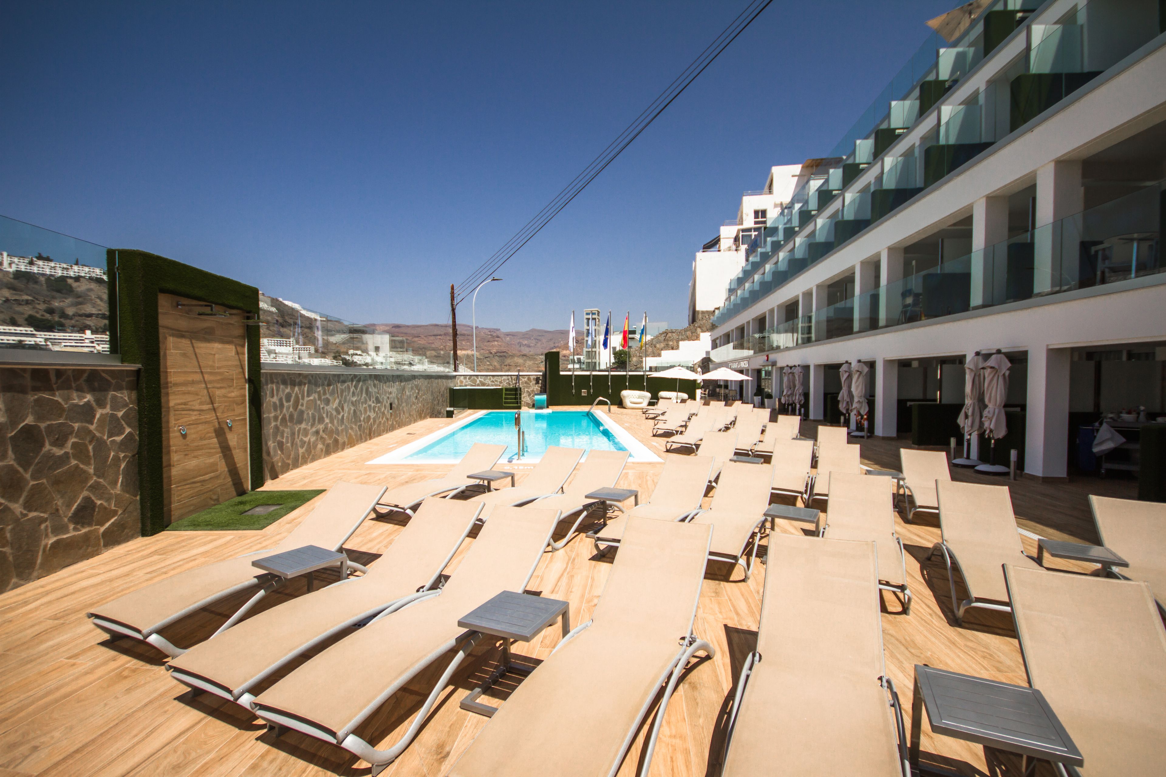 A poolside area with sun loungers, a swimming pool, and a view of a mountainous landscape.