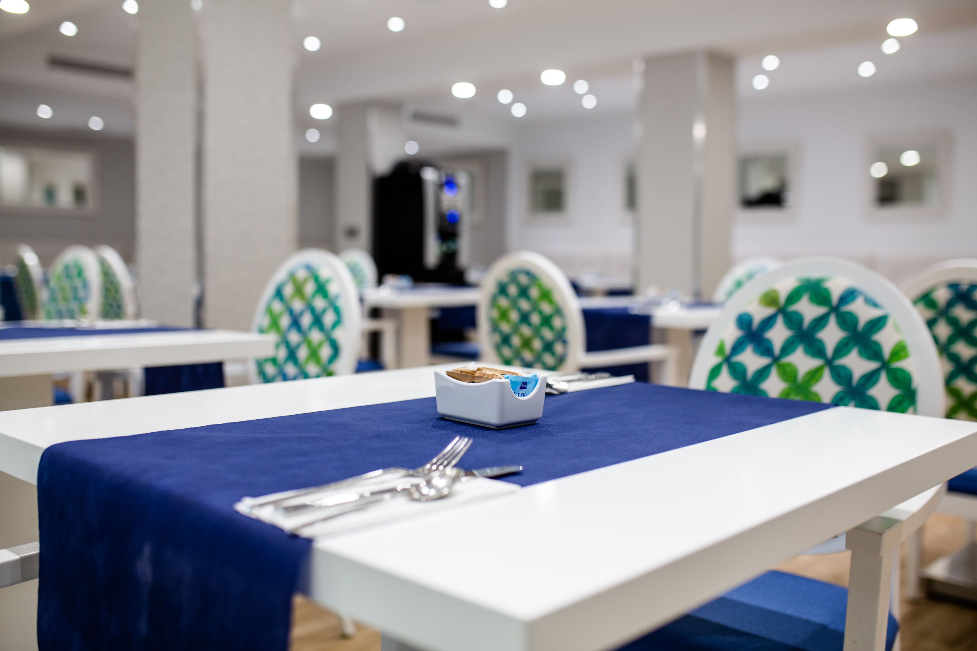A dining area with tables set for two, chairs with a geometric pattern, and a small white container with utensils on the table.