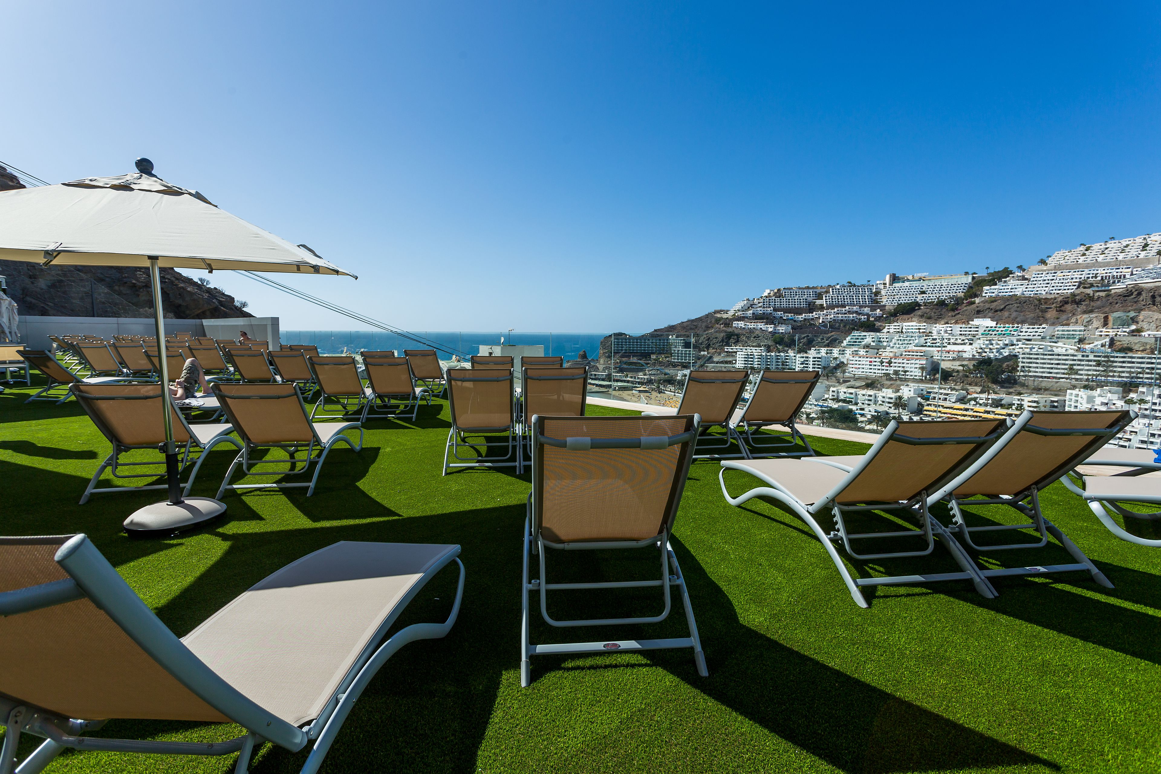 A rooftop terrace with rows of sun loungers and a large umbrella, overlooking a coastal cityscape.