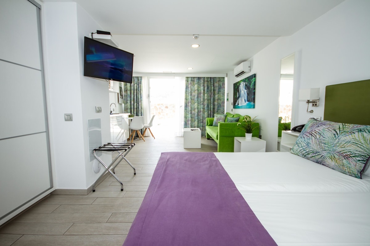 Una habitación de hotel moderna con una cama grande, un escritorio, un sofá verde y un televisor montado en la pared.
