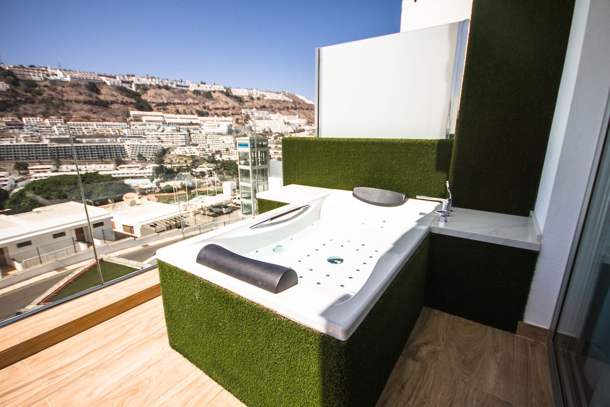 Un baño moderno con un detalle de césped artificial verde, una encimera blanca y vistas a un paisaje urbano.