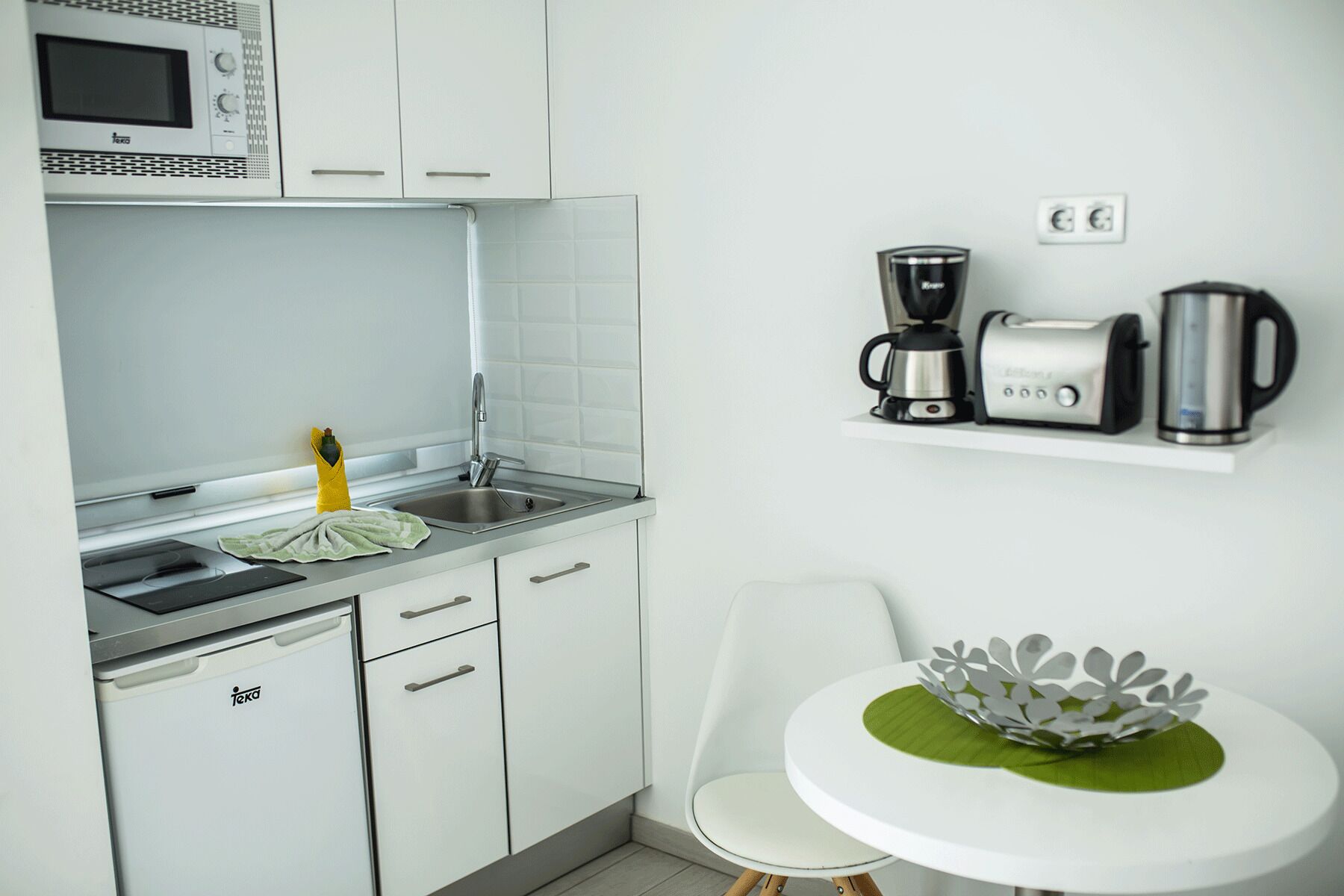 A compact kitchen with white cabinets, a microwave, a sink, and a small dining area with a white chair and a decorative bowl.