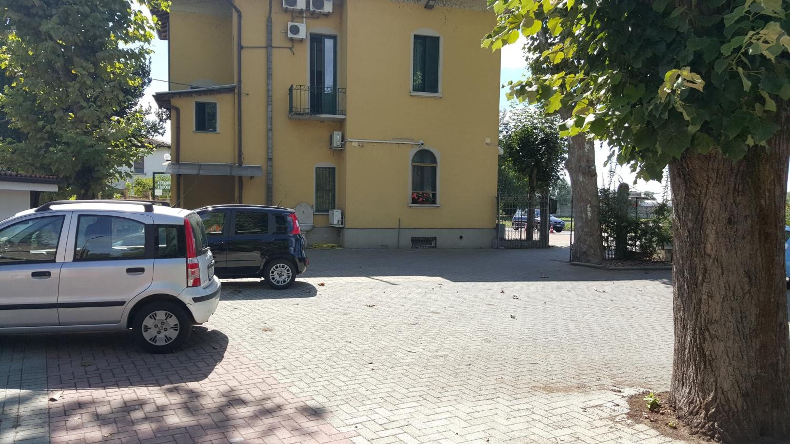 Eine gepflasterte Straße mit parkenden Autos, ein gelbes Gebäude mit mehreren Fenstern und Balkonen sowie ein Baum auf der rechten Seite.