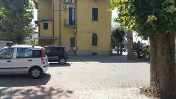 Eine gepflasterte Straße mit parkenden Autos, ein gelbes Gebäude mit mehreren Fenstern und Balkonen sowie ein Baum auf der rechten Seite.