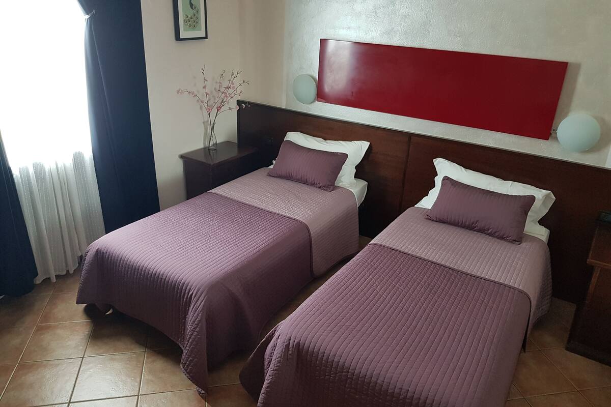A hotel room with two beds, each with a headboard and pillows, a nightstand, a vase with branches, and a red accent wall.