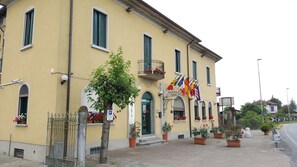A yellow building with a sign that reads "HOTEL La Penda." There are multiple flags, a tree, and potted plants in front of the building. - Hotel La Pendola (Ozzero)