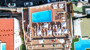 Vista aérea de un resort con una piscina central rodeada de sillas de sol y sombrillas, varias piscinas más pequeñas y un estacionamiento con coches.