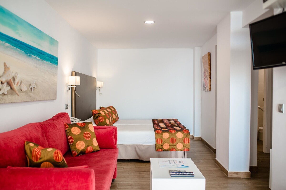 Habitación de hotel con un sofá rojo, una mesita de centro blanca, una cama con colcha estampada, un televisor montado en la pared y un cuadro con una escena de playa.