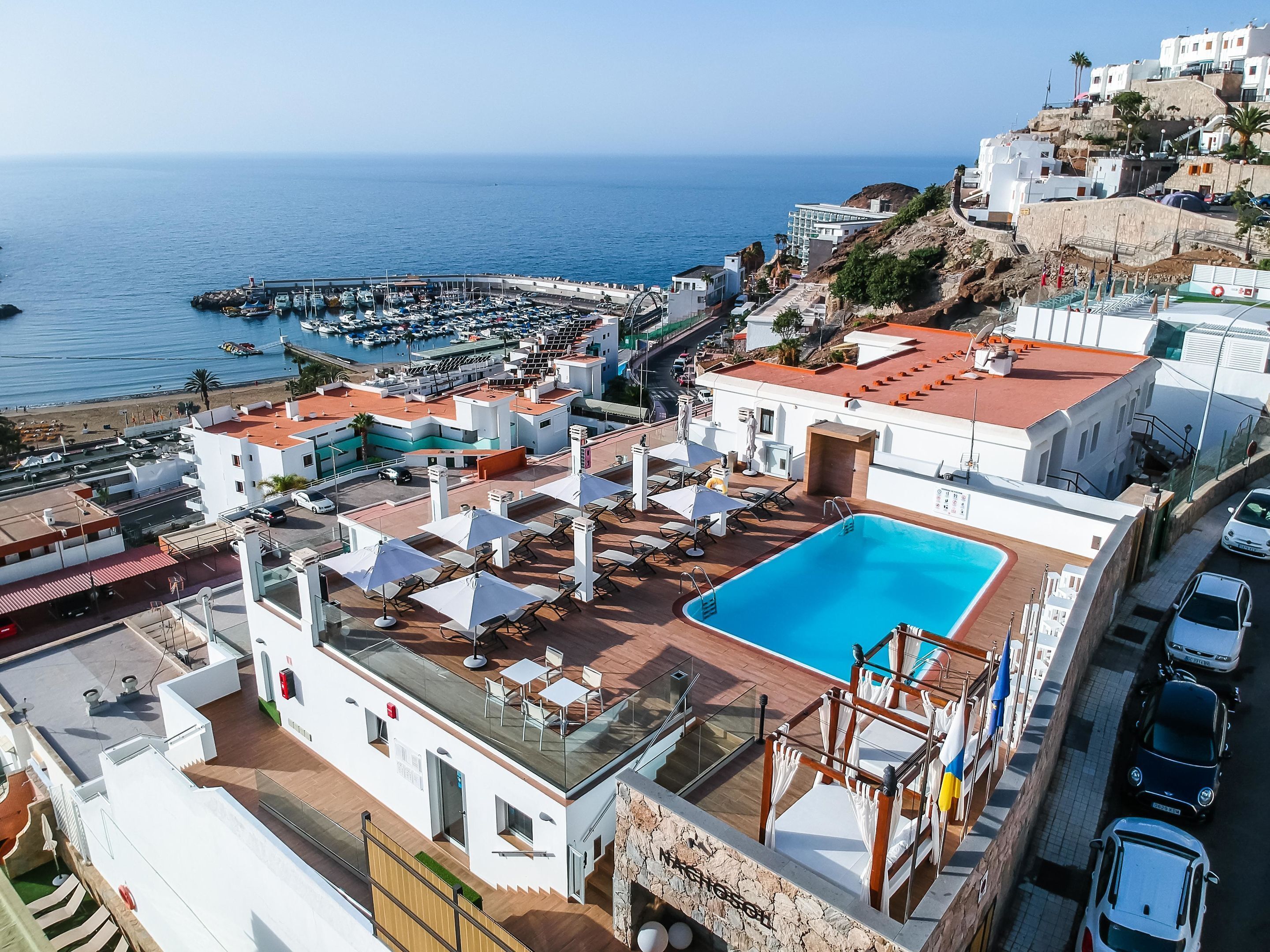 Area piscina sul tetto con vista libera sul mare, un porto turistico con barche e una collina con edifici.