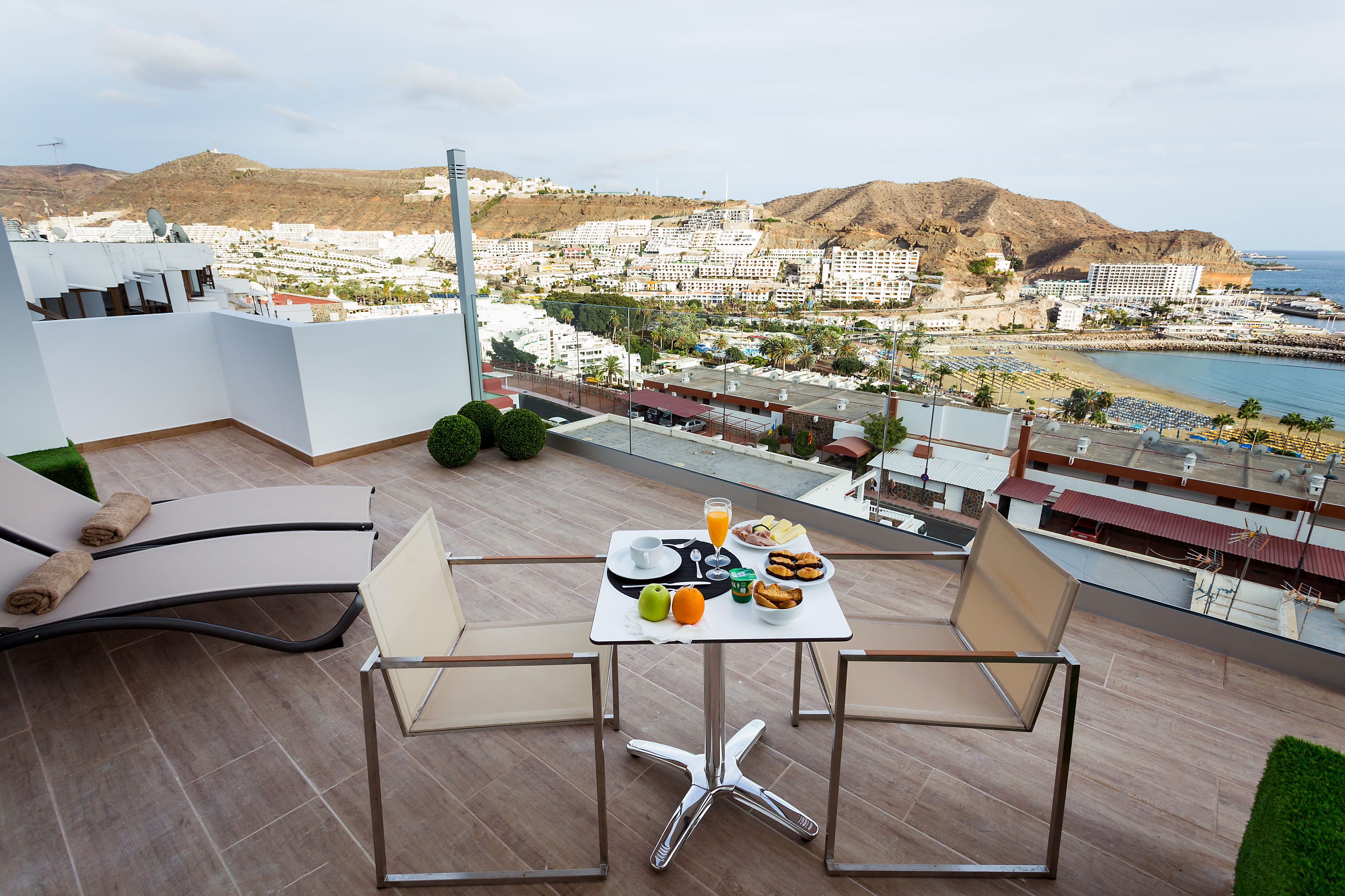 A balcony with a table set for breakfast, overlooking a coastal town and beach.