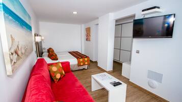 Una habitación de hotel moderna con un sofá rojo, una mesita blanca pequeña, un televisor montado en la pared y un cuadro con una playa.