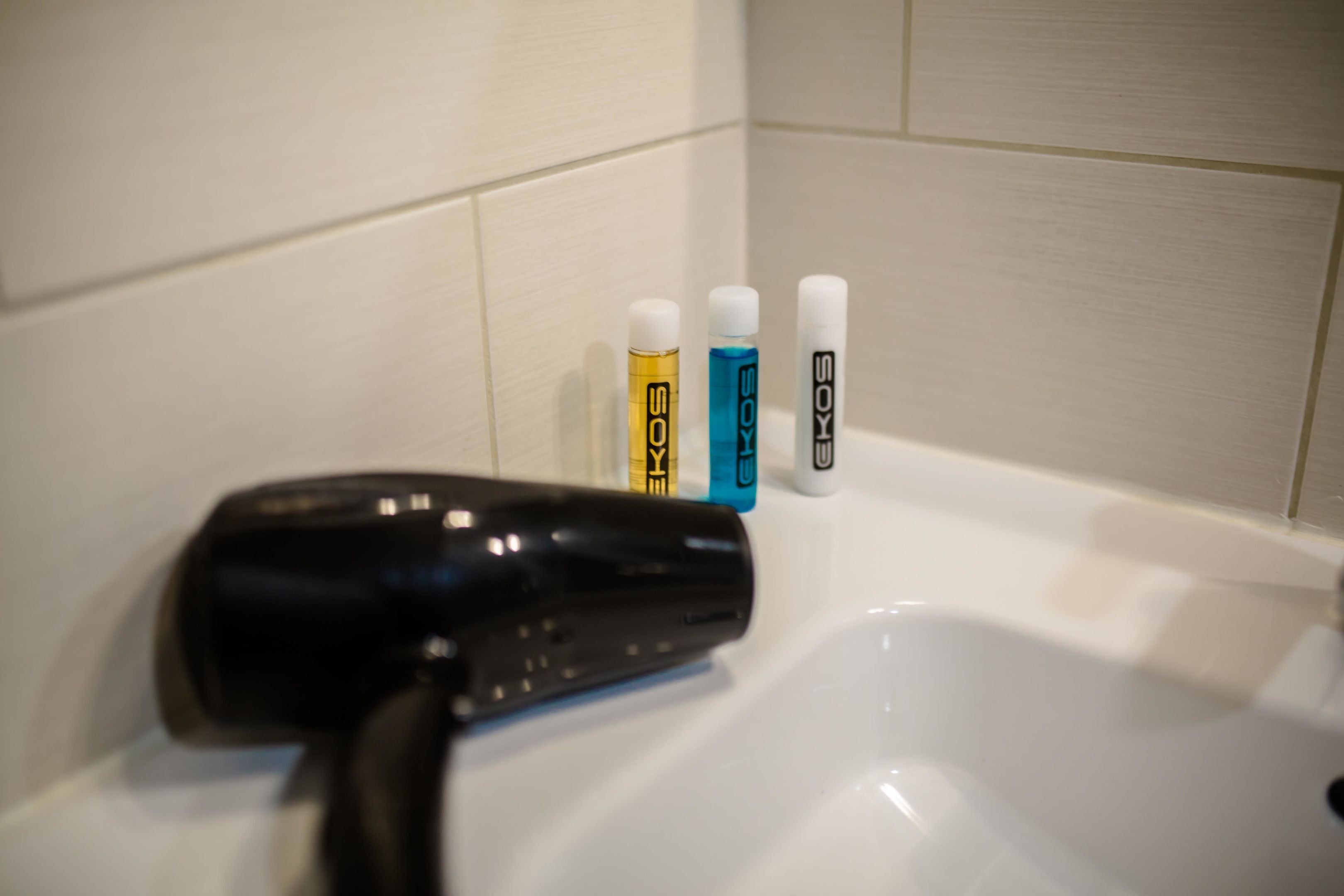 A bathroom with a black hairdryer on a sink, and three bottles of hair care products labeled 'SOKOS' in different colors.