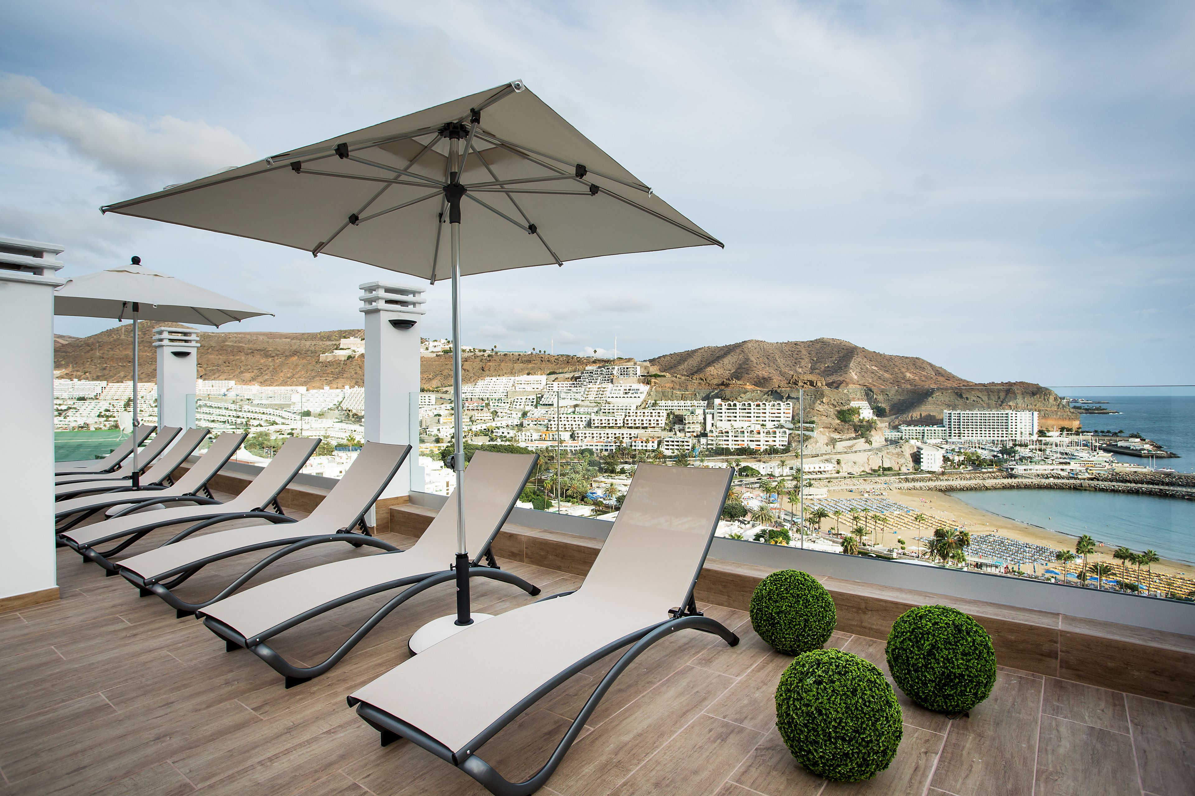 A balcony with sun loungers and a large umbrella, overlooking a coastal cityscape with buildings and a beach.