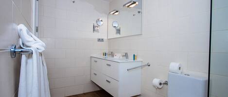Un baño moderno con azulejos blancos, suelo de madera, un lavamanos blanco con un espejo encima y un inodoro con un dispensador de toallas de papel.