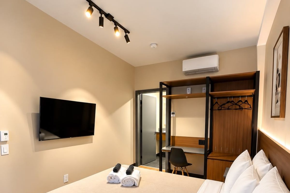 A hotel room with a large bed, a flat-screen TV mounted on the wall, a wooden wardrobe, and a desk with a chair.
