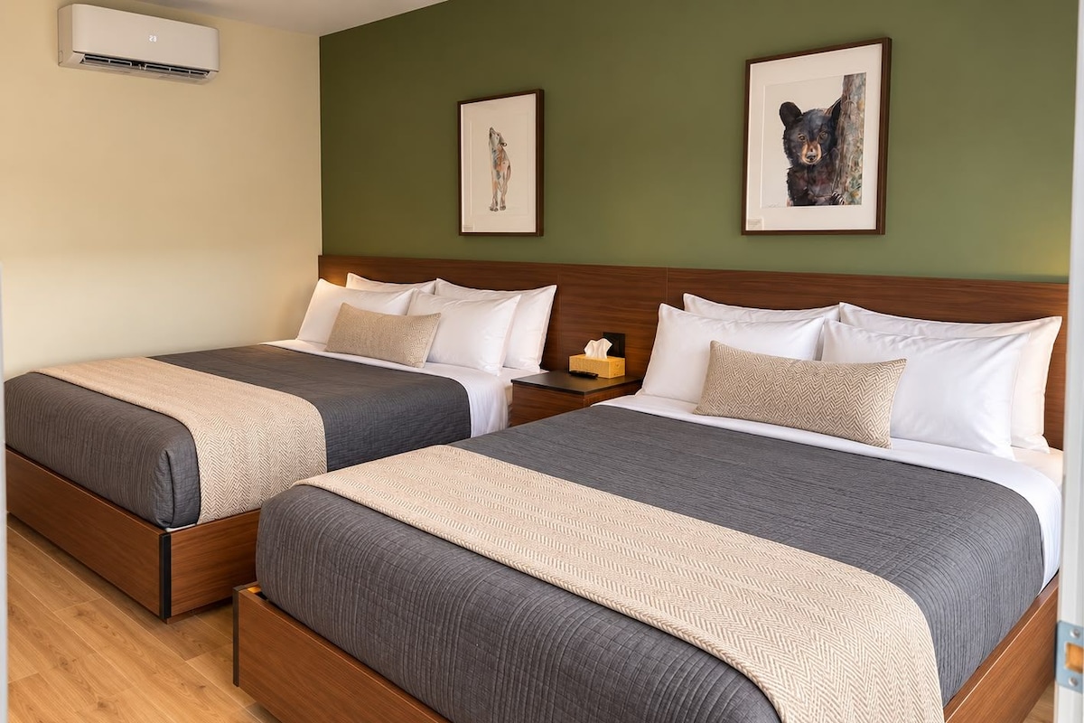A hotel room with two beds, a wooden headboard, a nightstand with a lamp, and framed artwork on the wall.