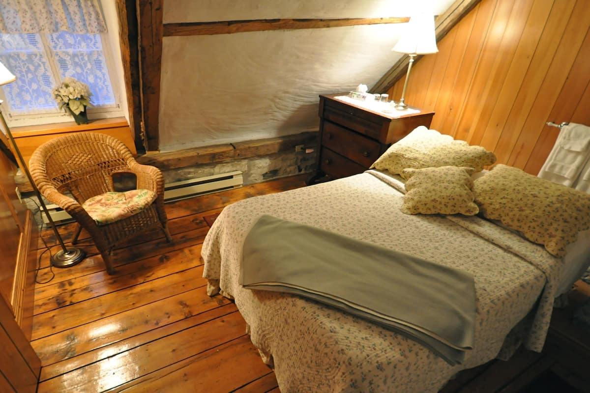 A cozy bedroom with wooden floors, a wicker chair, a bed with floral bedding, and a nightstand with a lamp.