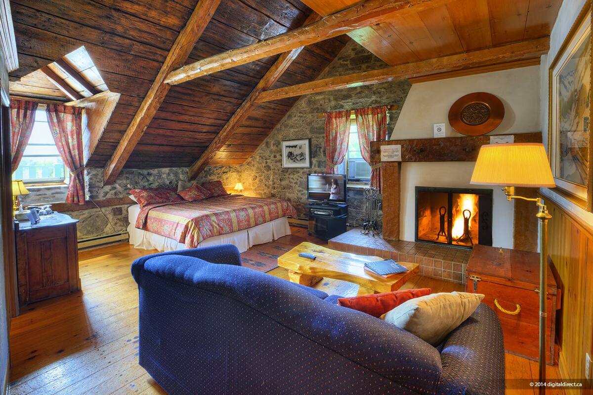A cozy attic bedroom with a wooden ceiling, a fireplace, a bed, a sofa, and a television.