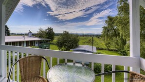 Een balkon met een rieten stoel en een glazen tafel, met uitzicht op een groen landschap met bomen en een huis.
