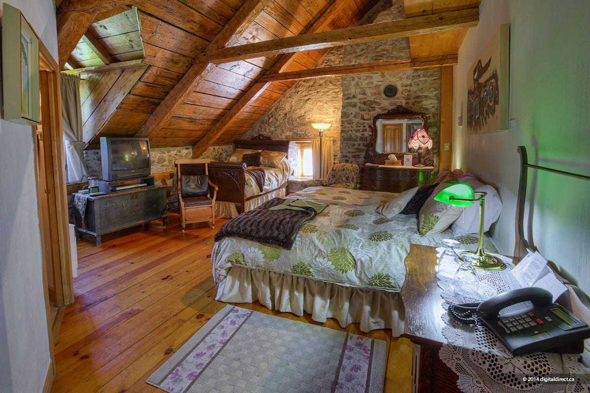 A cozy attic bedroom with a wooden ceiling, stone walls, a bed with a floral bedspread, a desk with a lamp, and a vintage television.