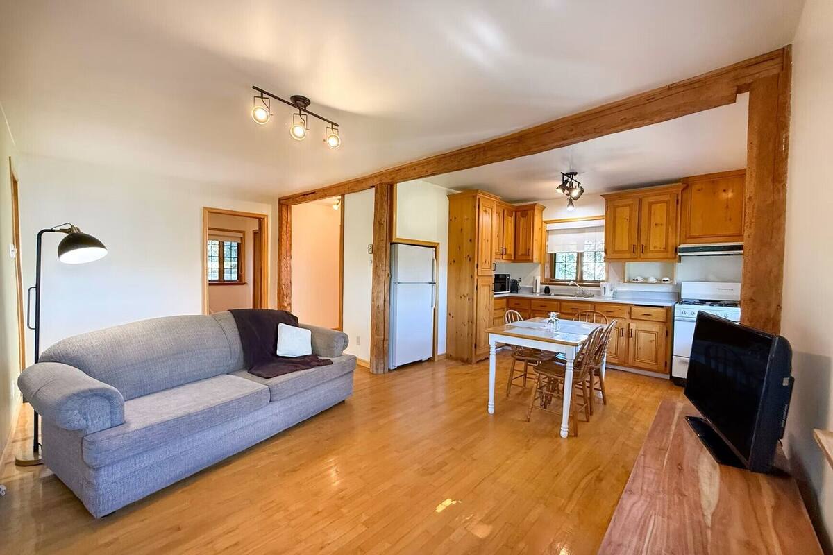 A living room with a sofa and a dining table, wooden floors, and a kitchen with wooden cabinets and appliances.