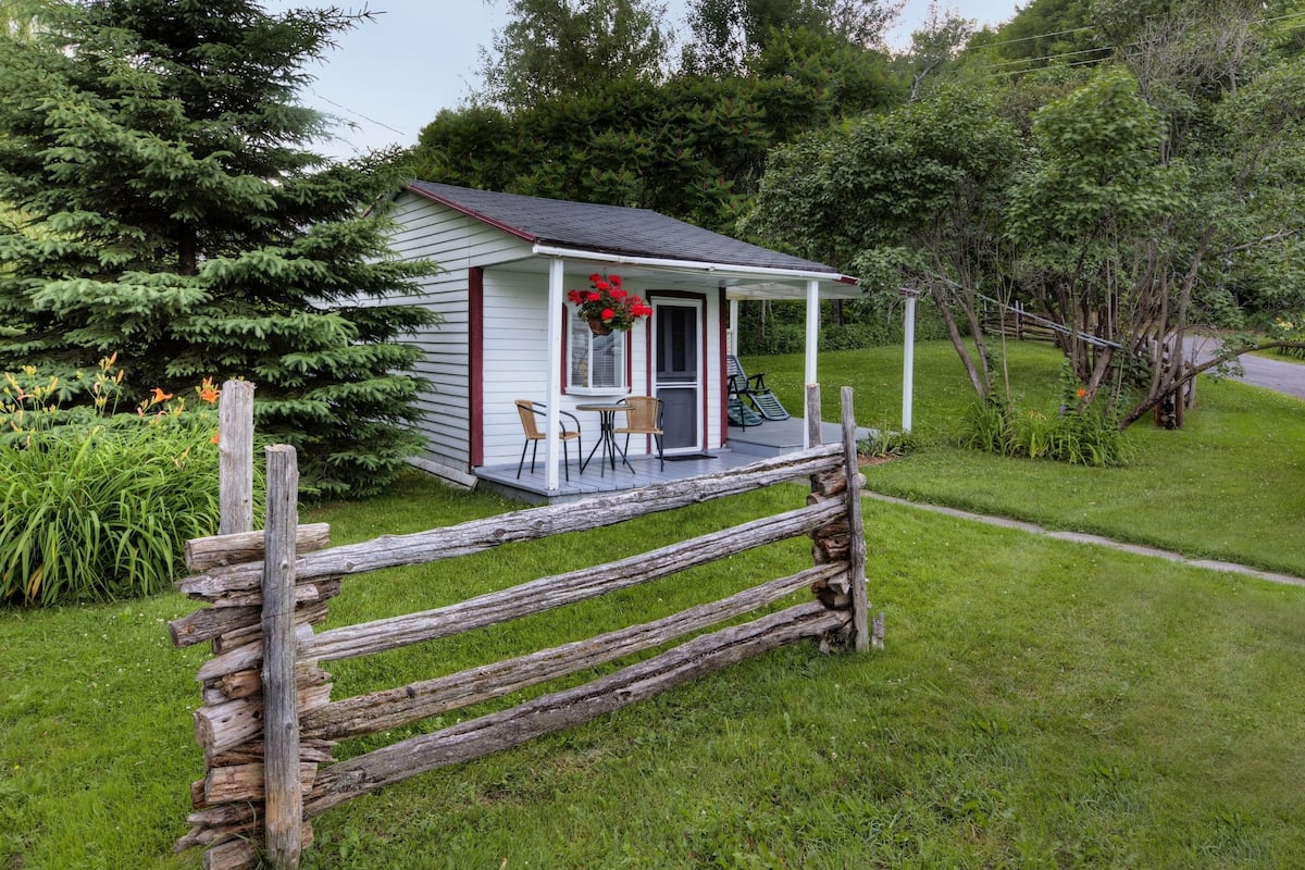 A small white cottage with a porch, surrounded by a wooden fence and lush greenery.