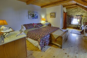 A bedroom with a wooden bed, a bedside table with a lamp, a small window with curtains, and a stone wall. - Auberge Baker (Chateau-Richer)