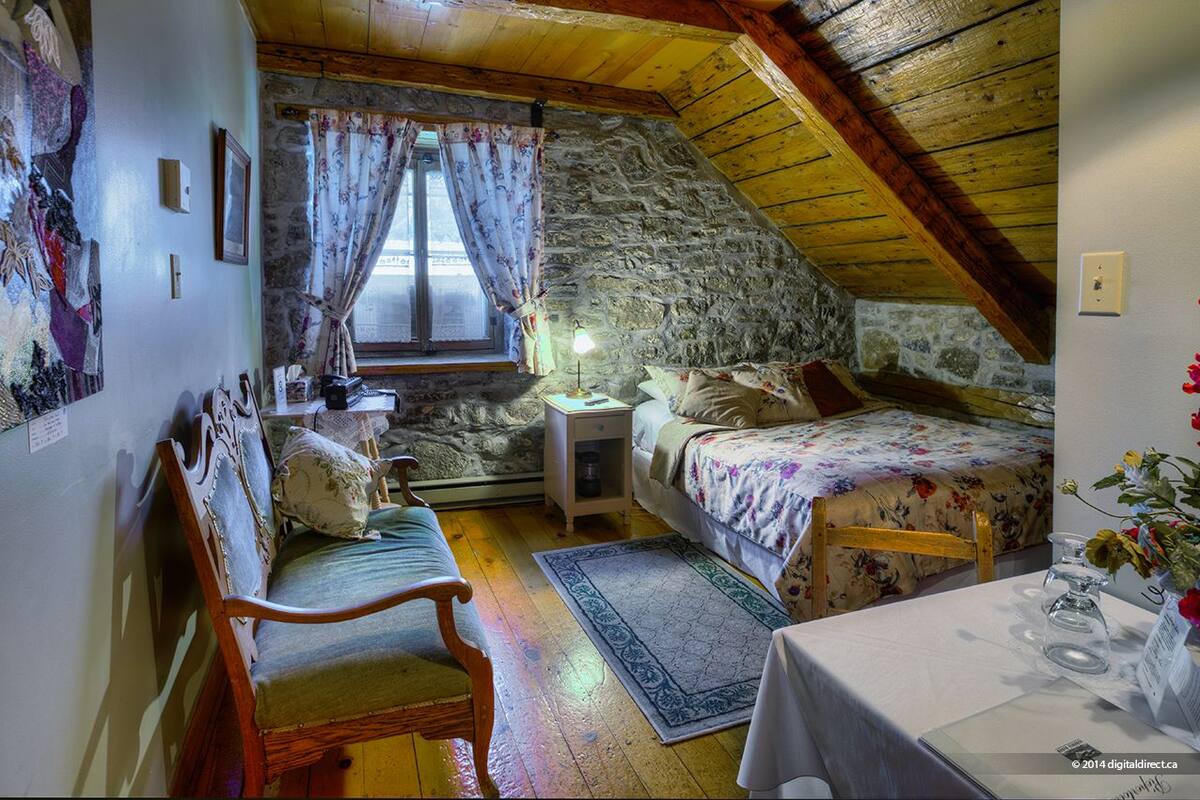 A cozy attic bedroom with a sloped ceiling, stone walls, a wooden bed, a wooden chair, a nightstand, and a window with floral curtains.
