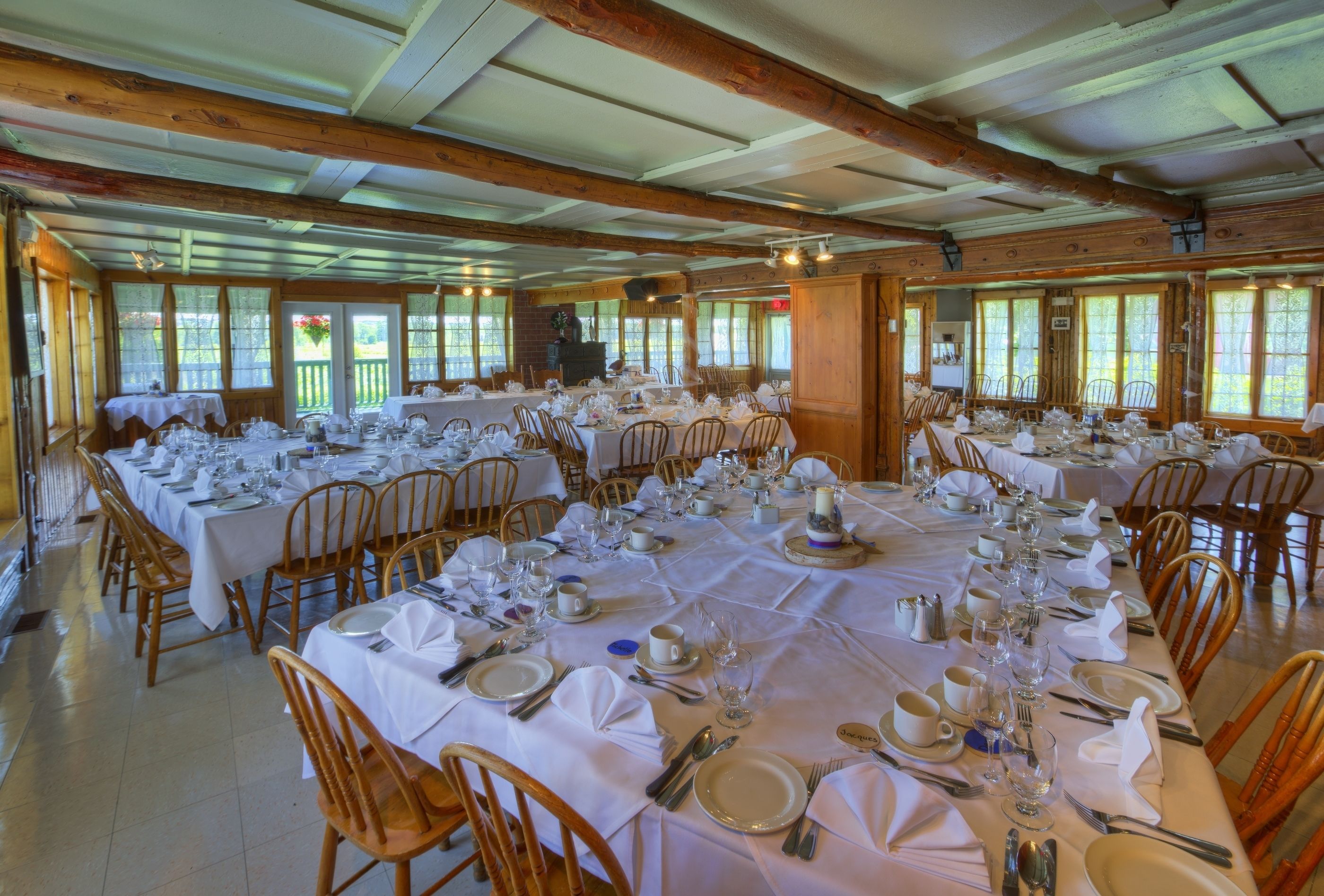 A large dining hall with multiple tables set for a formal event, wooden beams on the ceiling, and large windows allowing natural light.