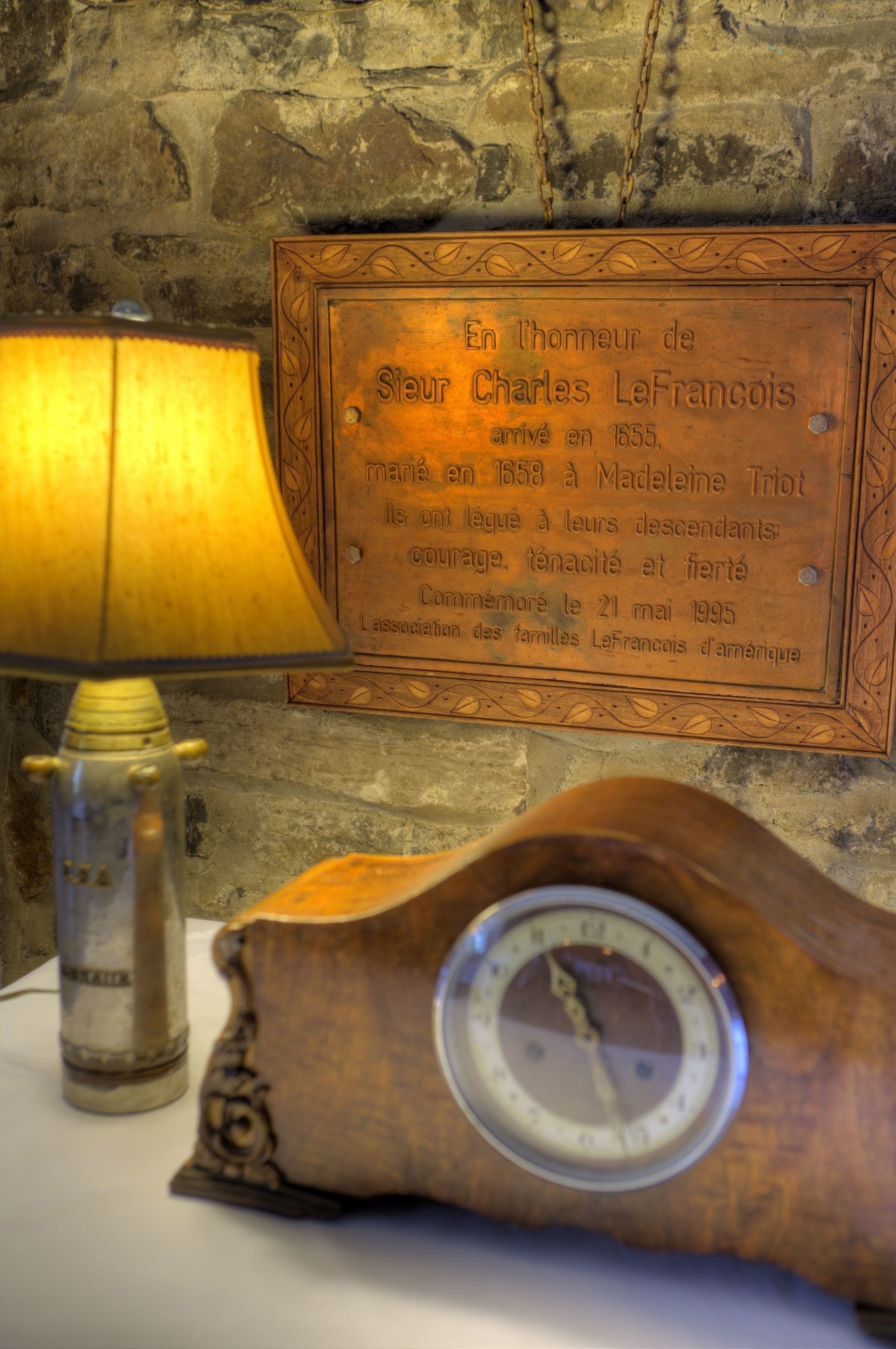 A wooden clock with a brass face, a brass lamp, and a plaque on a stone wall.
