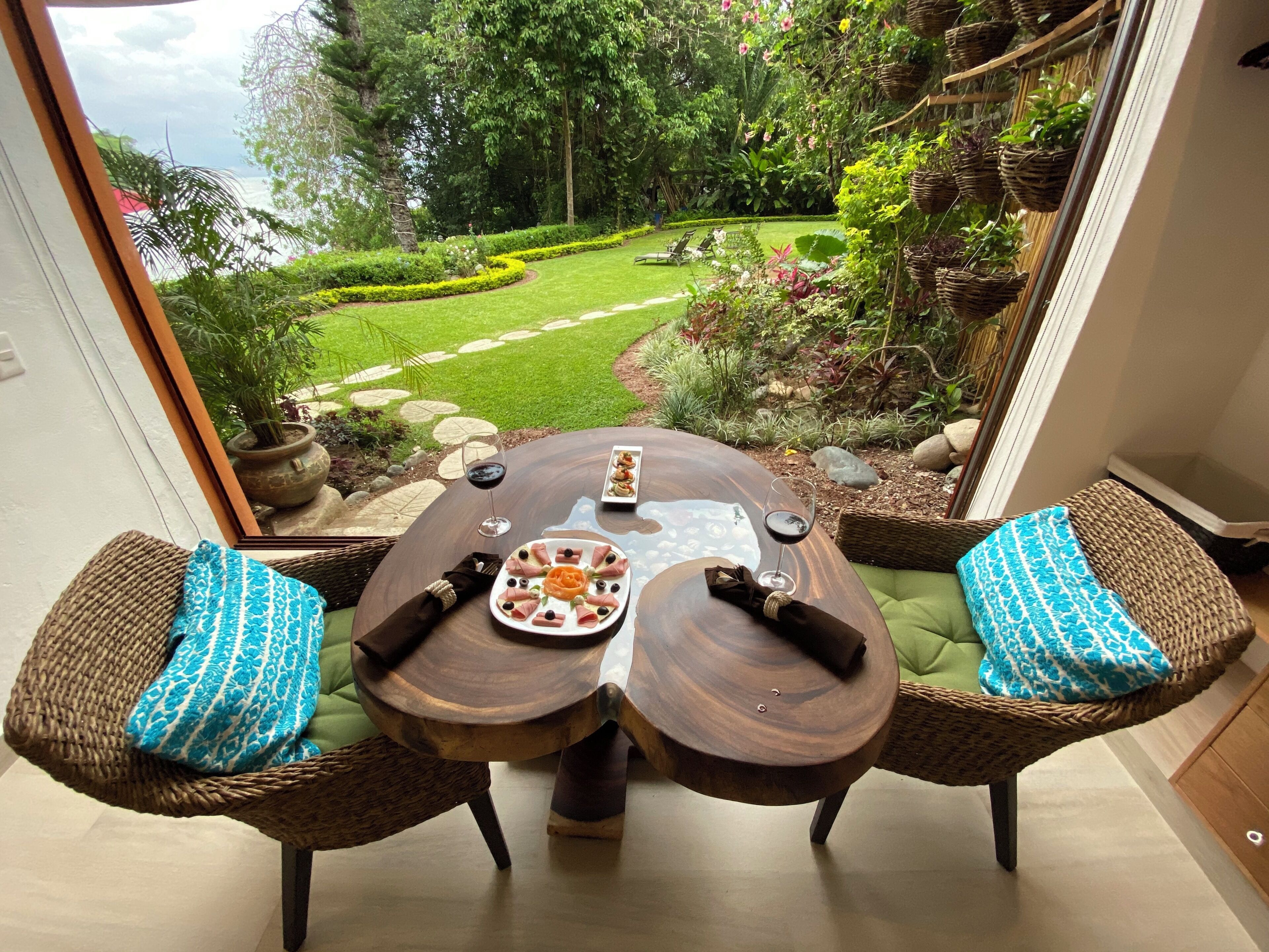 A dining table with two wicker chairs, a view of a garden with a lawn and trees, and a stone pathway.